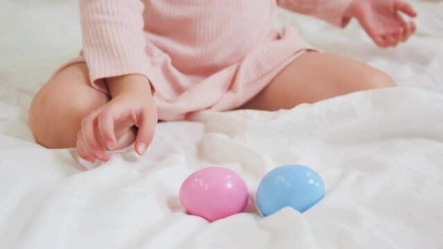 A girl of 4 or 5 years old in a pink dress is sitting on a white bed and playing with Easter eggs. A child rolls multicolored eggs on a white blanket. Close-up hands and two Easter eggs.