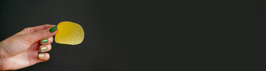 potato chips in woman's hand. girl holds light snack in her hand. black background. green manicure. Source of calories. horizontal image. Banner for insertion into site.