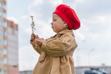 Charming Toddler in a beige raincoat and red beret admires a flower in spring.
