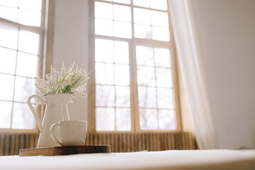 White vase with white small flowers on the table on the background of large windows in a wooden house