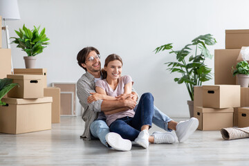 Glad millennial caucasian male in glasses and female in casual hugs, sits on floor among cardboard boxes