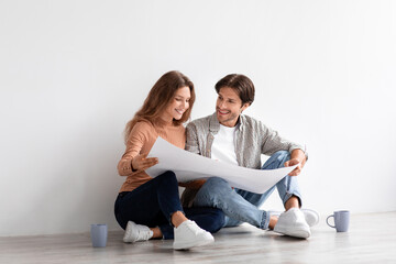 Fototapeta na wymiar Glad millennial caucasian male and female look at house plan sit on floor in empty room on white wall background