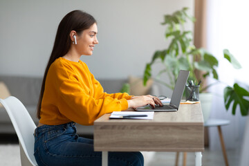 Cheery young woman in earphones using laptop, having online business meeting or educational webinar...
