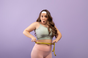 Fototapeta na wymiar Surprised plus size european millennial woman in sportswear with open mouth measures waist with measuring tape