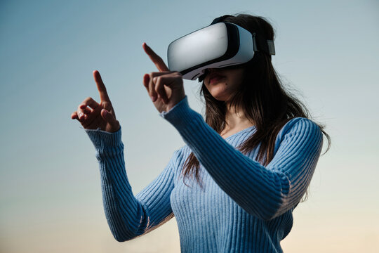 Young woman using virtual reality simulator gesturing in front of sunset sky