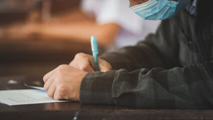 Student taking exam while wearing face mask due to coronavirus emergency. Young woman sitting in...