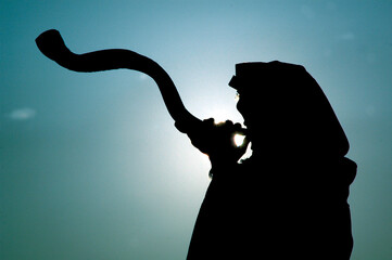 Silhouette of a Jewish man wearing a tallit and blowing a long, curly shofar made from the horn of...