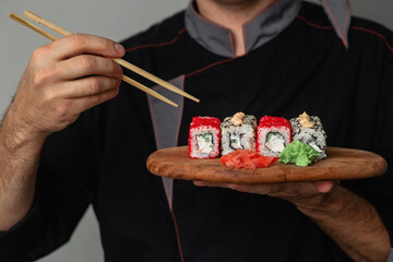 Close-up of the hands of a Man chef in a black uniform holding a wooden tray plate full of sushi...