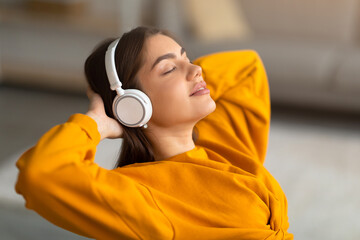 Calm young Caucasian woman listening to music in wireless headphones, relaxing with hands behind head at home