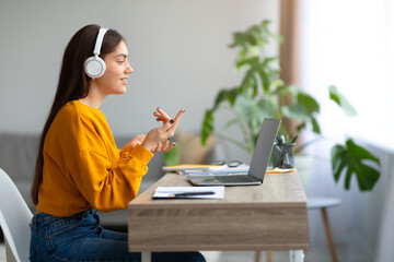 Happy young Caucasian female wearing headphones, using laptop, working or studying online from home, empty space