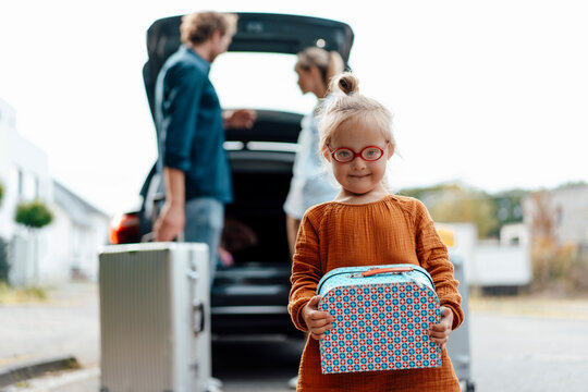 Girl holding suitcase with parents loading luggage in car