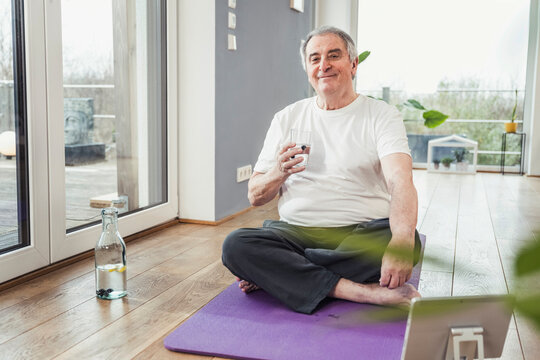 Smiling senior man sitting cross-legged with drinking glass on exercise mat at home