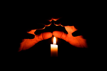 Hands folded over the flame of a candle on a black background. The concept is a talisman, the...