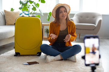 Young travel vlogger recording video content on smartphone, sitting on floor near suitcase,...