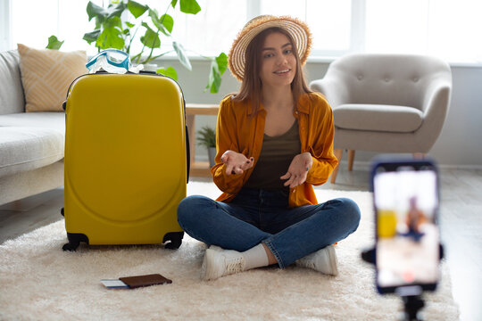 Young travel vlogger recording video content on smartphone, sitting on floor near suitcase, speaking to blog followers