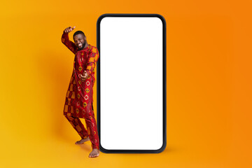 Black man in traditional costume dancing and having fun near big smartphone with empty screen, orange background