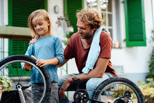 Smiling Father Looking At Son Cleaning Bicycle Outside House