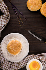 bread concept buns, a dish of flour, a knife, and a tiny bowl of raw egg lying on the dark wooden table