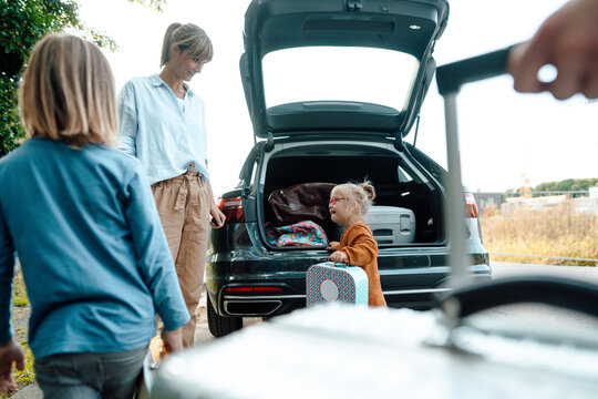 Mother looking at daughter with suitcase by car trunk on vacation
