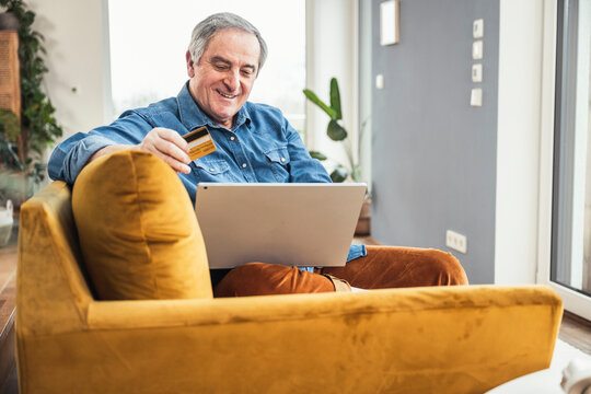 Smiling senior man with laptop and credit card sitting on sofa in living room