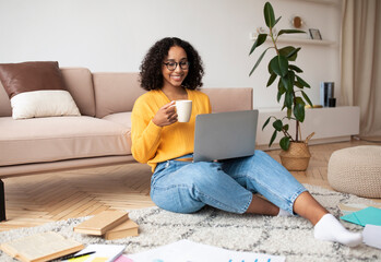 Cheery young black woman having video call with family or friend on laptop, drinking coffee at home, copy space