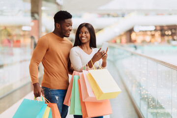 Portrait of excited black spouses using phone holding shopping bags