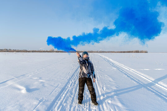 A male teenager in winter in an open space holds a smoke bomb with blue smoke in his hands. The concept of freedom and rebellion.