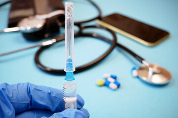 syringe in doctor's hand in medical gloves, pills and stethoscope on smartphone on blue background, concept of telemedicine or e-health