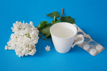Pills from seasonal spring allergy. Cup of water, pill, lilac flowers on blue background. Pollen allergies concept