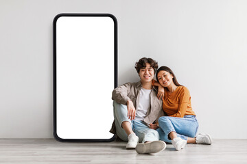 Loving asian couple sitting near big cellphone with empty white screen, embracing, recommending app...