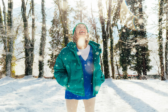 Cheerful Woman With Hands In Pockets Enjoying Snowfall At Forest