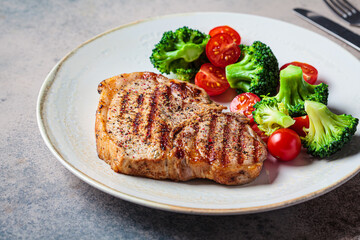 Grilled pork steak with broccoli and tomatoes on plate, dark background.