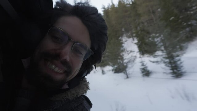 Close-up of young man face with beard and long hair and glasses smiling enjoying his train ride through snowy natural landscape, sustainable green travel concept hanging outside window