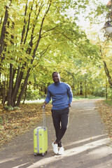 Cheerful african american young man in stylish clothes with suitcase walks in an spring park on warm sunny day. Countryside travel concept