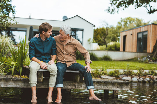 Senior man talking with son sitting on jetty by lake at backyard