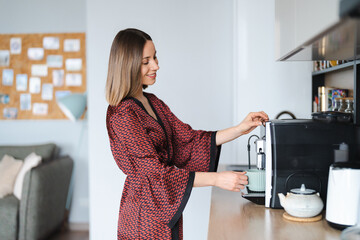 Woman using coffee machine to make big mug of coffee at home. Woman wearing silk robe at home while...