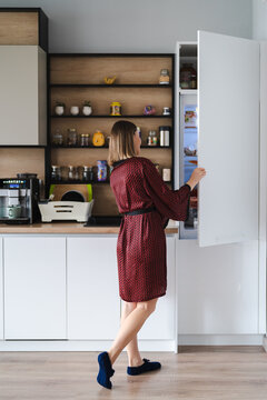 Hungry Woman Looking For Food In the Fridge At Home, but don't have Much there. White kitchen furniture, home wear, red silk robe