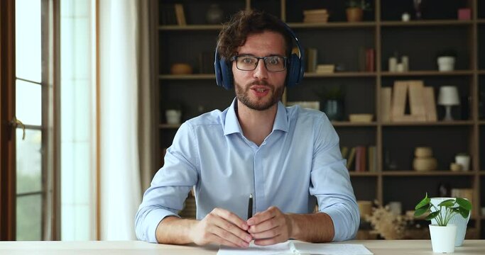 Smiling confident millennial male teacher in headset sit at office desk give remote video lecture talk to students before web camera. Young man in glasses speaking at webcam conference videocall event