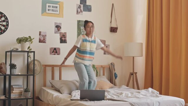 Full-length shot of gen z Asian teenage girl in knitted vest and blue jeans jumping on bed at home