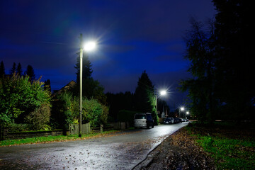 Abgelegene n&auml;chtliche Strassenszene im l&auml;ndlichen Raum mit Beleuchtung. Im Hintergrund sieht man parkende Autos