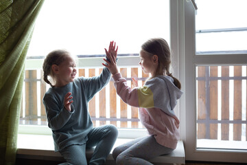 two girls are playing patty-cake, sitting on the windowsill near the big window