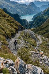 Alpine landscape with a path winding through the mountains and a lake in the distance. A girl is walking on the path, with her back to the photographer and heading towards the lake.
