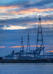 Fototapeta na wymiar Riverbank with large electricity masts and container cranes at twilight, Port of Antwerp.