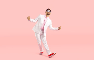 Happy funny guy in white suit dancing isolated on pastel pink background. Full body shot of cheerful goofy carefree man in modern suit, sneakers and glasses dancing and having fun in the studio