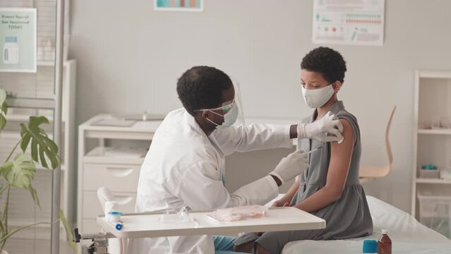 Medium slowmo shot of 10-year-old African-American girl receiving Covid-19 vaccine at modern doctors office