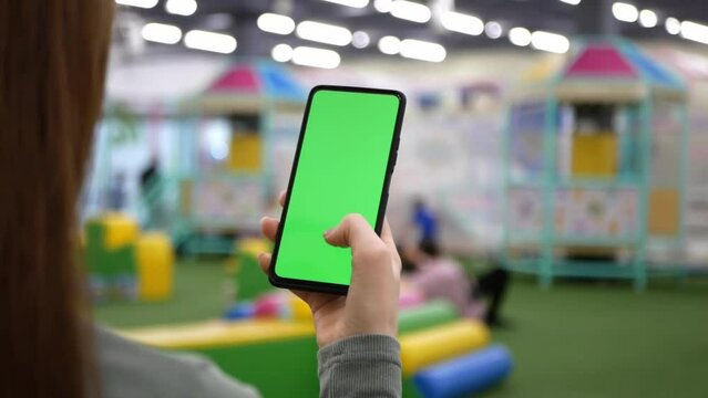 Young mother uses smartphone with green mock-up screen. Concept of Internet surfing in public place. Woman taps green mockup screen while waits children in entertainment or game center.