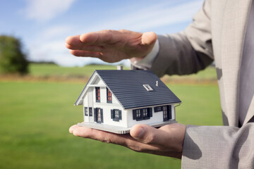Hand holding a model house in empty green grass field covering and protecting insurance security concept