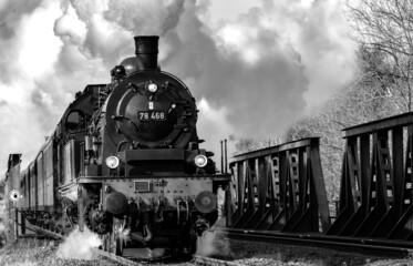 Steam train with historic locomotive and coaches crossing steel rivet bridge in Ruhr valley between Arnsberg and Meschede Sauerland Germany. Historic railway black and white greyscale vintage.