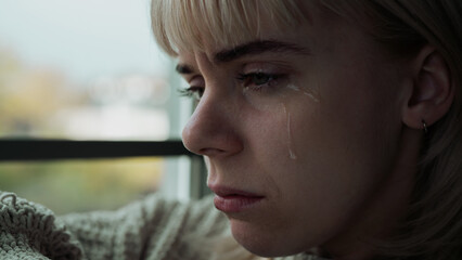 Close Up of a Beautiful Blonde and Young Caucasian Woman Feeling Depressed and Crying While Looking Down. Tears Falling and Light Shining on Females Face From a Big Window.
