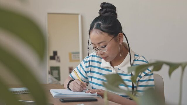 Waist-up shot of gen z Asian teenage girl writing in copybook then taking her smartphone starting scrolling sitting at desk in her room